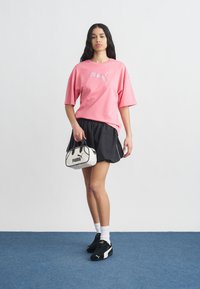 Femme debout, portant un T-shirt rose Puma, un short noir, des baskets noires Puma, des chaussettes blanches, et tenant un sac à main blanc Puma.