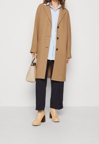 Camel uldblandet frakke med store revers, knaplukning, stylet med en blå skjorte, marineblå bukser og beige støvler med hæl.