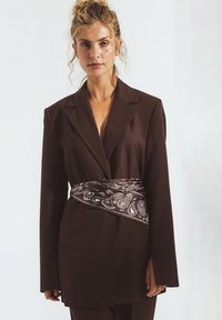 Brauner maßgeschneiderter Blazer mit langen Ärmeln, der mit einem am Bauch gebundenen Paisley-muster Scarf versehen ist. Glattes Material, strukturiertes Design.