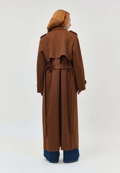 Persona con lunghi capelli rossi che indossa un lungo trench marrone con cintura e jeans blu a gamba larga, con le spalle rivolte verso un fondo semplice.