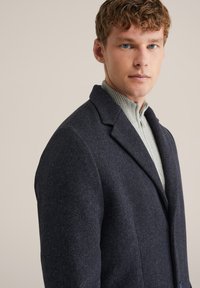 Cappotto di lana blu scuro con revers strutturati, design monopetto e texture morbida. Indossato sopra un maglione a collo a coste grigio chiaro.