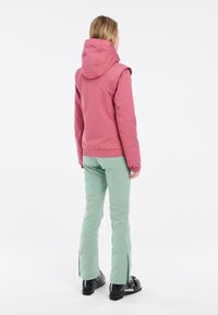Rosa isolierte Jacke mit Kapuze und elastischen Bündchen, kombiniert mit grünen Skihosen mit seitlichen Reißverschlüssen. Schwarze Skistiefel vervollständigen das Outfit.