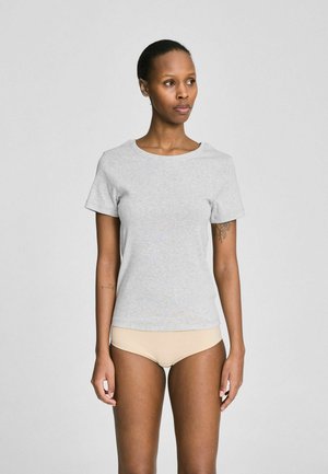 Donna che indossa una T-shirt grigio chiaro a maniche corte e biancheria intima beige, in piedi contro uno sfondo grigio semplice, che guarda verso destra.