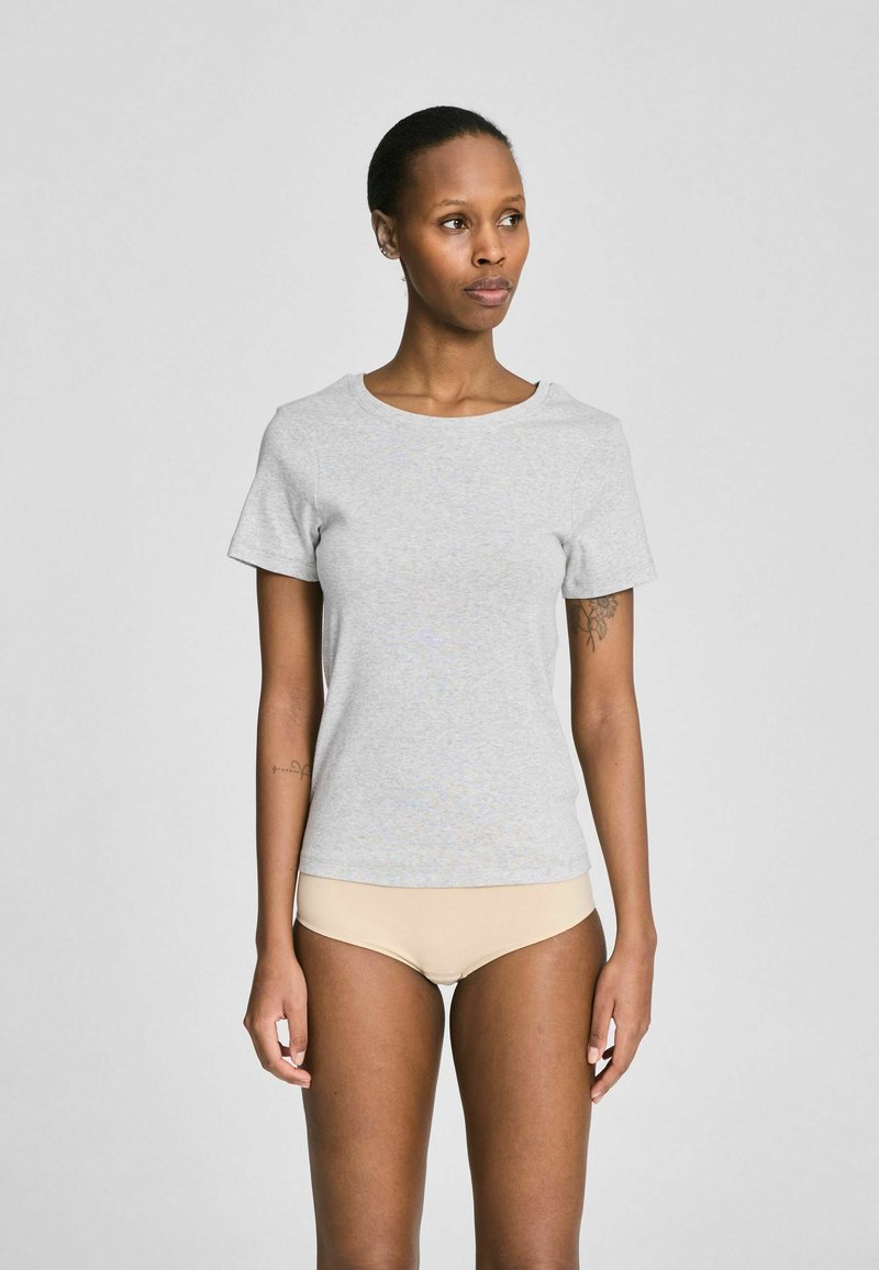 Žena u svijetlosivoj kratkorukavnoj majici i bež donjem rublju stoji nasuprot jednobojnoj sivoj pozadini, gleda prema desno.