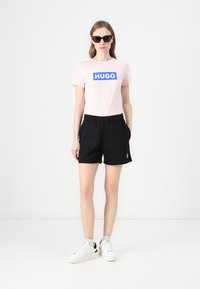 Pink cotton t-shirt with blue "HUGO" logo, paired with black shorts and white sneakers. Sunglasses complete the casual look.