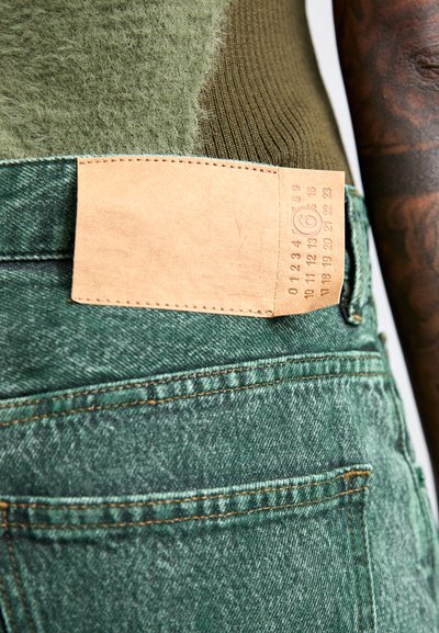 Πράσινα τζιν με υφή, διαθέτοντας ένα ανοιχτό καφέ δερμάτινο λεκέ με ανάγλυφες λεπτομέρειες κατά μήκος της ζώνης.