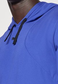 Felpa con cappuccio in un vivace tessuto blu, caratterizzata da una superficie testurizzata, lacci neri e un piccolo logo nero vicino al collo.