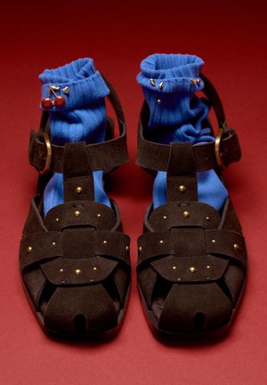 Paar dunkelbraune Ledersandalen mit goldfarbenen Nieten, getragen mit leuchtend blauen, gerippten Socken, die mit kleinen Broschen auf rotem Hintergrund verziert sind.