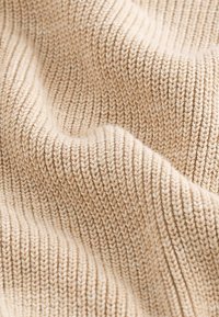Beige kootud kangas ribade tekstuuriga, näidates pehmet ja paindlikku materjali koos nähtavate õmbluste ja looduslike kiudude detailidega.