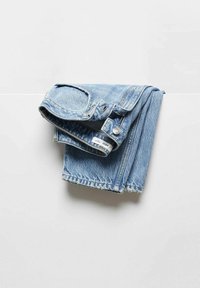 Jean en denim bleu clair plié et suspendu sur une surface horizontale blanche sur un fond pâle.