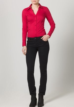Frau trägt ein eng anliegendes rotes Hemd mit langen Ärmeln und Knöpfen, schwarze Skinny-Jeans und schwarze Knöchel-Stiefel, steht mit einer Hand in der Tasche.