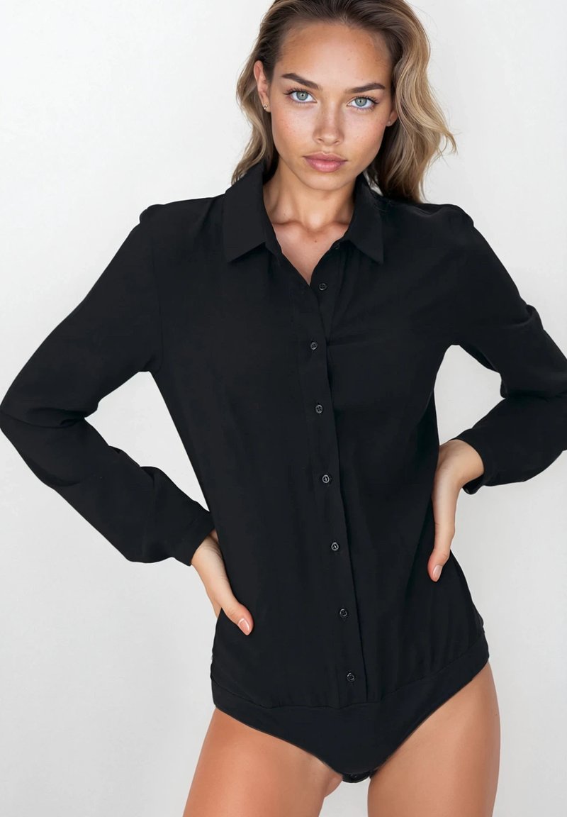 Kobieta o jasnych włosach, ubrana w czarny, długi rękaw bodysuit zapinany na guziki, stojąca z rękami na biodrach na jednolitym białym tle.