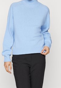 Pull tricoté bleu clair avec un col montant, des poignets et un ourlet côtelés, présentant une coupe décontractée et une texture douce. Associé à un pantalon noir.