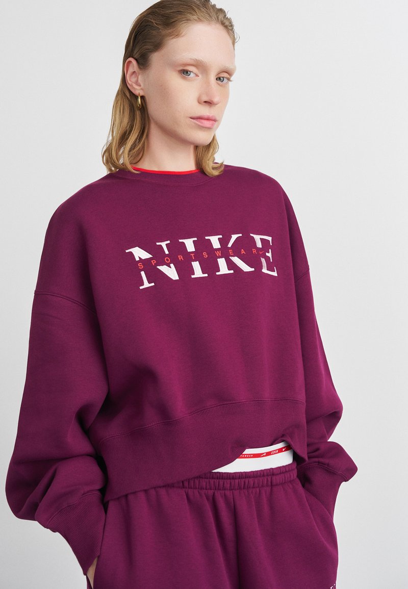 Giovane donna che indossa una felpa Nike viola e pantaloni jogger abbinati, con le mani in tasca su uno sfondo semplice.