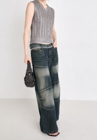 Haut sans manches gris en maille torsadée, associé à un jean en denim large et déchiré avec une texture unique et une ceinture chaîne. Le mannequin tient un sac en perles.