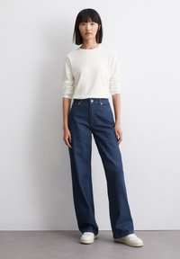 Hvid langærmet skjorte og høj talje mørkeblå bred ben jeans. Sneakers med en struktureret hvid sål komplementerer outfittet.