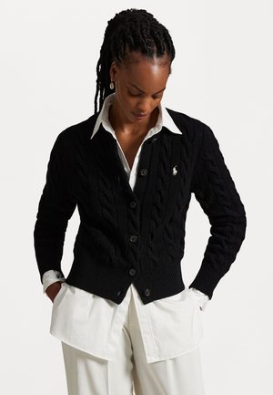 Woman with braided hair wearing a black cable-knit cardigan over a white collared shirt and loose white pants, hands in pockets, looking down.