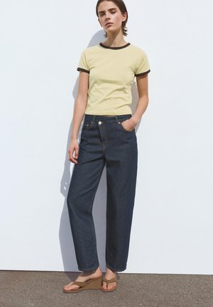 Junge Frau steht vor einer weißen Wand, trägt ein gelbes Ringer-T-Shirt, dunkle, hoch taillierte, weit geschnittene Jeans und braune Keilsandalen.