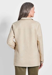 Veste en faux daim beige avec un col, des manches longues et des poignets boutonnés. Présente une coupe droite et un détail de couture visible au dos.
