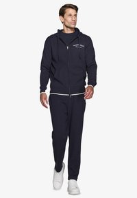 Marineblå zip-up hoodie med en kengurulomme, let stof, kombineret med matchende bukser og hvide sneakers. Har snoretræk ved hætten.