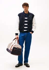 Veste varsity navy et blanche avec un t-shirt rayé, jeans bleu et chaussures noires ; tenant un sac de sport beige et navy avec des accents marron.