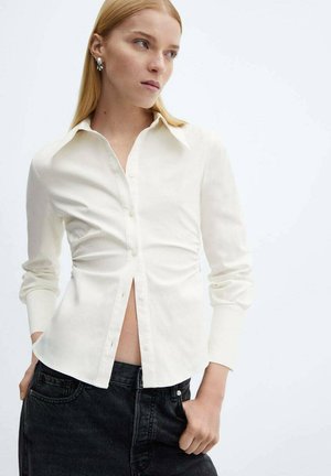 Chemise blanche à manches longues avec un col, coupe ajustée, boutonnière à l'avant et plis discrets sur les côtés ; assortie à un jean foncé.
