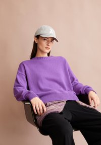 Junge Frau, die eine graue Baseballkappe und einen lila Pullover trägt, sitzt entspannt in einem Stuhl vor einem beigen Hintergrund.