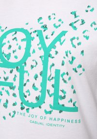 Weißes T-Shirt mit grünem Text, der "JOYFUL" liest, versehen mit einem Muster aus verstreuten grünen Leopardenflecken und kleinerem Text darunter.