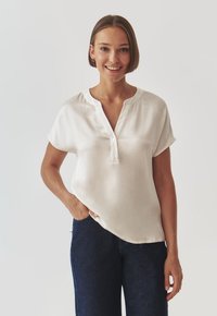 Short-sleeve, off-white blouse with a notch collar, featuring a button placket. Smooth texture, relaxed fit, paired with dark denim jeans.