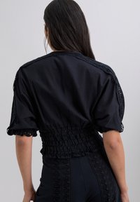 Blouse en coton noir avec des manches courtes bouffantes, taille smockée et des détails en dentelle élaborés le long du col et des manches. Texture lisse.