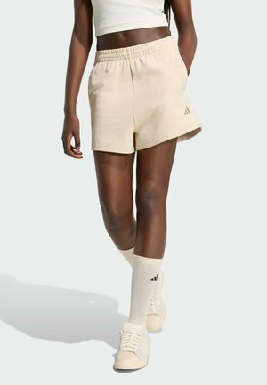 Persona snella che indossa pantaloncini sportivi beige, calzini alti abbinati, scarpe da ginnastica bianche e un top corto bianco su uno sfondo semplice.