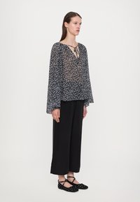 Blouse noire en dentelle florale avec des manches longues en forme de cloche, décolleté en V et détail à nouer. Associée à un pantalon large noir et des chaussures noires à enfiler avec des clous.