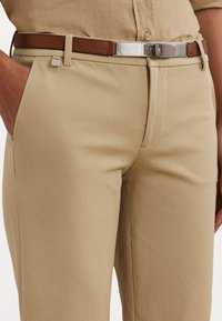 Beige skräddarsydda byxor med en slät textur. Kombinerat med ett brunt läderbälte med en silverfärgade spänne och en fyrkantig accentdetalj.