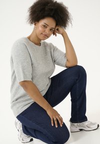 Světle šedý pletený svetr s krátkým rukávem, zkombinovaný s tmavě modrými džíny. Bílé a šedé sportovní boty. Modelka klečí a opírá ruku o koleno.