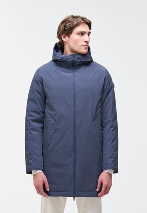 Hombre que lleva una chaqueta azul acolchada hasta la rodilla con capucha, de pie y mirando hacia un lado contra un fondo liso.