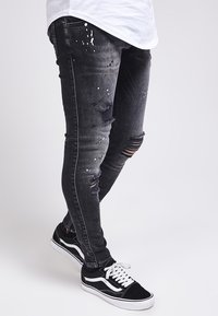 Zwarte gescheurde jeans met verfspatten, slim fit, met verschillende versleten plekken. Gecombineerd met zwart-witte sneakers.