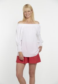 Weiße schulterfreie Bluse mit langen Ärmeln und ausgestellten Manschetten, kombiniert mit leuchtend roten Shorts aus leichten Materialien.