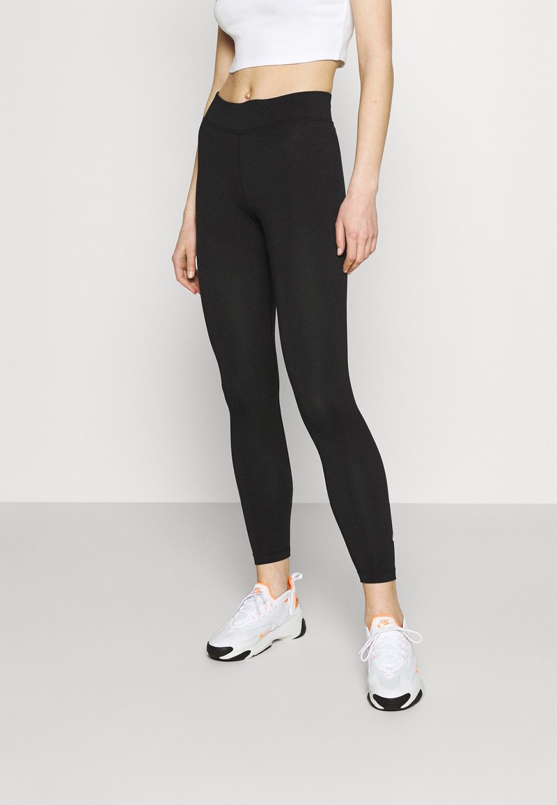 Svarta stretchiga leggings med hög midja, figurnära design, slät textur, kombinerat med vita sneakers med orange detaljer.