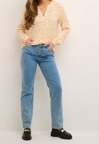 Blusa amarilla con estampado, mangas largas, escote en V profundo, combinada con jeans de tiro alto azul claro y zapatos negros gruesos con detalles en dorado.