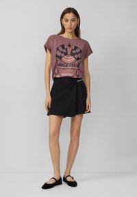 Bordeauxrotes Graphic-T-Shirt mit "Smile Club"-Aufdruck, kombiniert mit einem schwarzen Denim-Rock. Schwarze Slipper runden das Outfit ab.