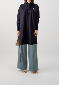 Marinebluse mit Knopfverschluss, langen Ärmeln und gesticktem Logo, kombiniert mit weit geschnittenen, hellblauen Jeans und beigen Sandalen.