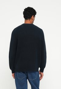 Maglione a coste blu scuro con maniche lunghe, scollo rotondo e una vestibilità morbida. La texture del tessuto appare soffice con un motivo a maglia robusto.
