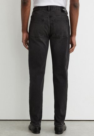 Bărbat purtând blugi negri cu picior drept și pantofi negri, stând în fața unui perete alb, vedere din spate cu buzunare și etichetă la curea.