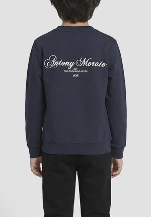 Kind draagt marineblauwe trui met de tekst "Antony Morato Est. Two Thousand Seven AM" op de achterkant, gecombineerd met zwarte broek, met de rug naar ons toe.