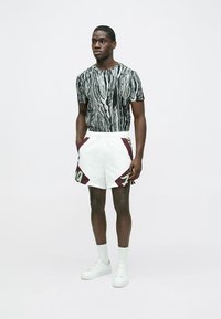 Melnbalta rakstaina īsām piedurknēm T-krekls, kopā ar baltām šortām ar tumši sarkaniem akcentiem un zaļiem burtiem. Balti sporta apavi.