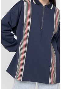 Suéter navy con cuello de media cremallera, que presenta rayas verticales en rojo, verde y beige. Fabricado en tejido suave, con un corte relajado y mangas largas.