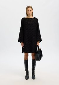 Svart texturerad klänning med lös passform och vida ärmar, kombinerad med knähöga svarta läderstövlar och en strukturerad svart handväska.