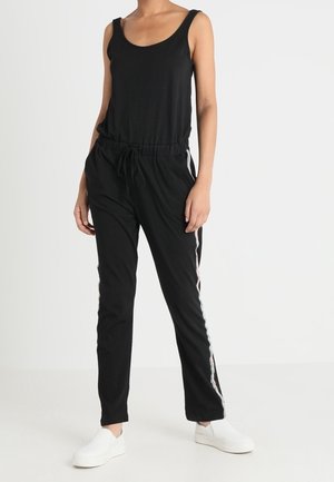 Personne portant une combinaison noire sans manches avec des bandes latérales et des chaussures blanches sans lacets, debout les mains dans les poches devant un fond blanc.