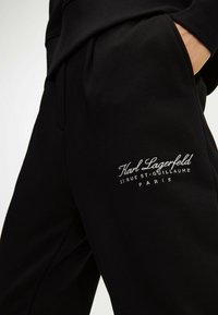 Black pants with hand in pocket and white embroidered text "Karl Lagerfeld 21 Rue St-Guillaume Paris" on thigh.