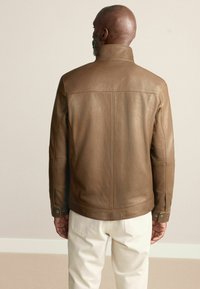 Jachetă din piele maro cu guler înalt, cusături structurate și mansete cu capse, asortată cu pantaloni de culoare deschisă. Textură netedă.